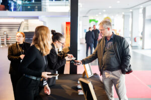 A person in a black jacket and glasses hands a device to a staff member at a counter in a modern indoor space.