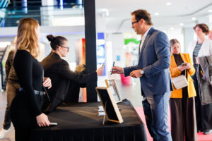 A group of people at an event interact near a table with devices and framed displays, suggesting a tech or business demonstration.