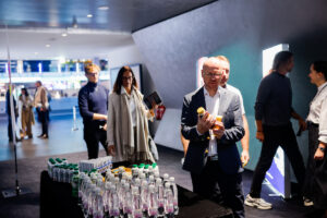A group of people in a modern indoor space gather around a table with bottled water and beverages, with one individual examining a product closely.