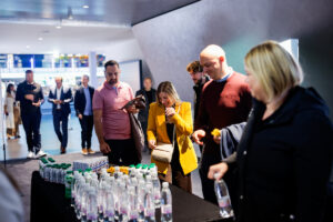 A group of people gather around a table filled with bottled water and snacks in a modern indoor setting.