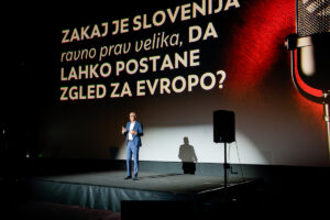 A person stands on a dimly lit stage in front of a large screen displaying a Slovenian question about Slovenia’s potential as a European prospect.