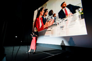 A presenter stands on a dimly lit stage in front of a large screen displaying a video conference with multiple participants.