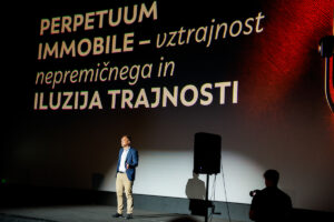 A person stands on a stage in front of a large screen displaying text in Slovenian about perpetuum immobile and the illusion of permanence.