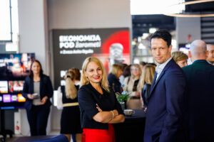 Two individuals pose confidently at a professional event, with a backdrop displaying "EKONOMIJA POD ŽAROMETI" and other attendees visible in the background.
