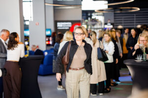 A person with short blonde hair and glasses walks through a modern event space filled with attendees and stylish furniture.
