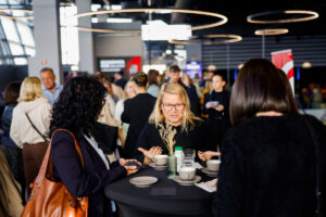 Three individuals engage in conversation around a table at a bustling indoor event, with modern circular lighting overhead.