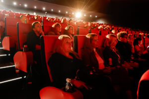 Audience members sit in a dimly lit theater with red seats, attentively watching a presentation under a bright spotlight.
