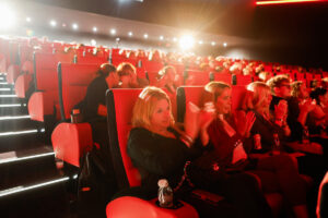 Audience members in a dimly lit theater with red seats applaud during a performance.