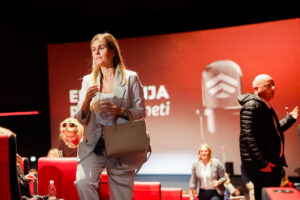 A person in a light-colored suit walks across a stage with red seating and a large screen displaying text and a logo.