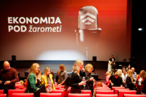 Audience members sit in red seats at an event with a large screen displaying "EKONOMIJA POD žarometi" and a microphone graphic.
