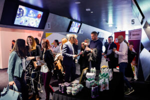 A group of people socialize at a modern event space with digital screens, branded signage, and a table stocked with beverages.