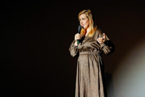 A person with long blonde hair stands on a dark stage, holding a microphone and gesturing while speaking.