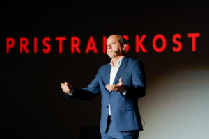 A person in a blue suit stands on stage speaking into a microphone, with the word "PRISTRANSKOST" displayed in red letters behind them.