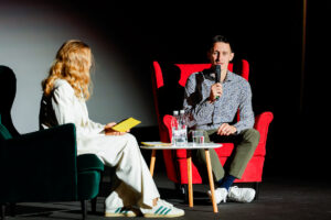 Two individuals engage in a conversation on stage, with one holding a microphone and the other seated nearby with a yellow notepad.