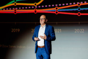 A person in a blue suit stands on stage, gesturing while speaking, with a colorful graph displaying data from 2019 to 2022 behind them.