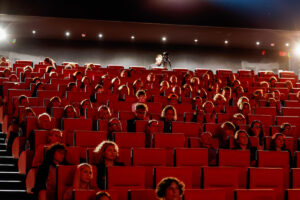 Audience members seated in a dimly lit theater with red chairs, attentively watching a screen, while a person with a camera stands at the back.